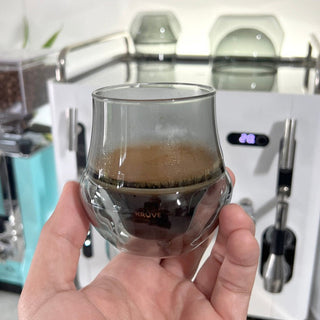 Hand holding KRUVE PROPEL Espresso Glasses (75ml/2.5oz) (2-Pack) with espresso inside and coffee machine background